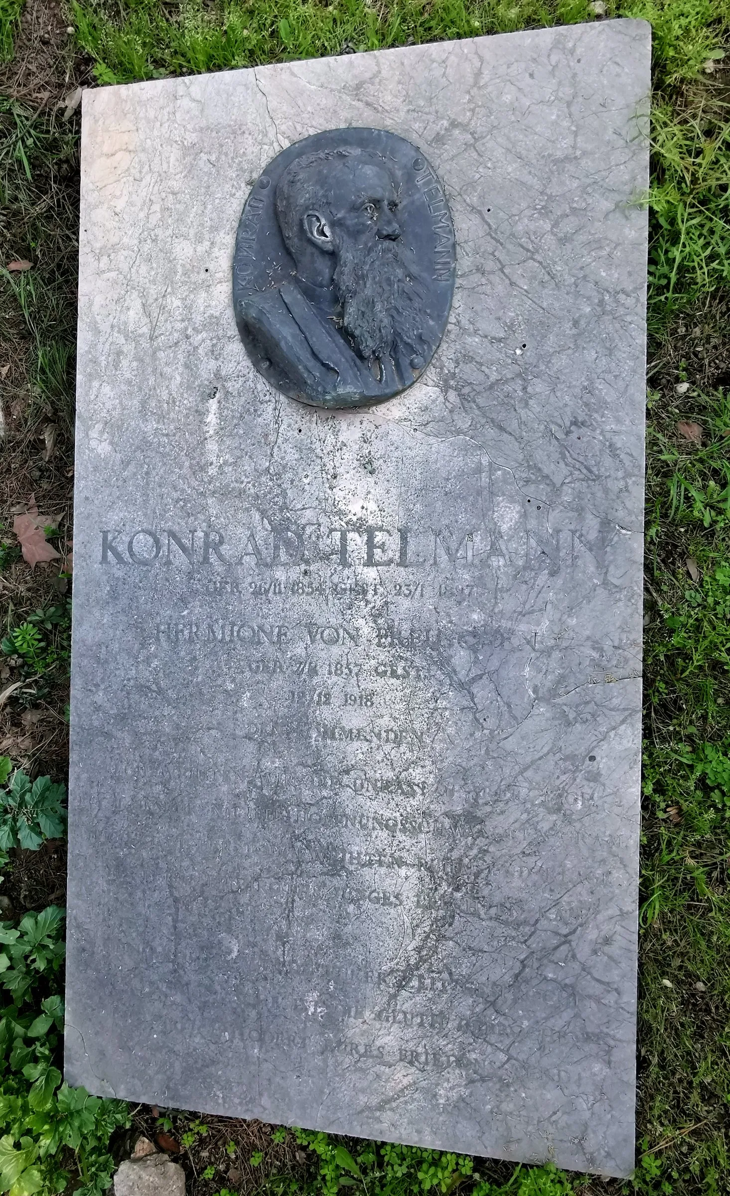 A flat marble slab set in grass, weathered and stained, carrying a bronze relief portrait of a bearded man and a long worn inscription.