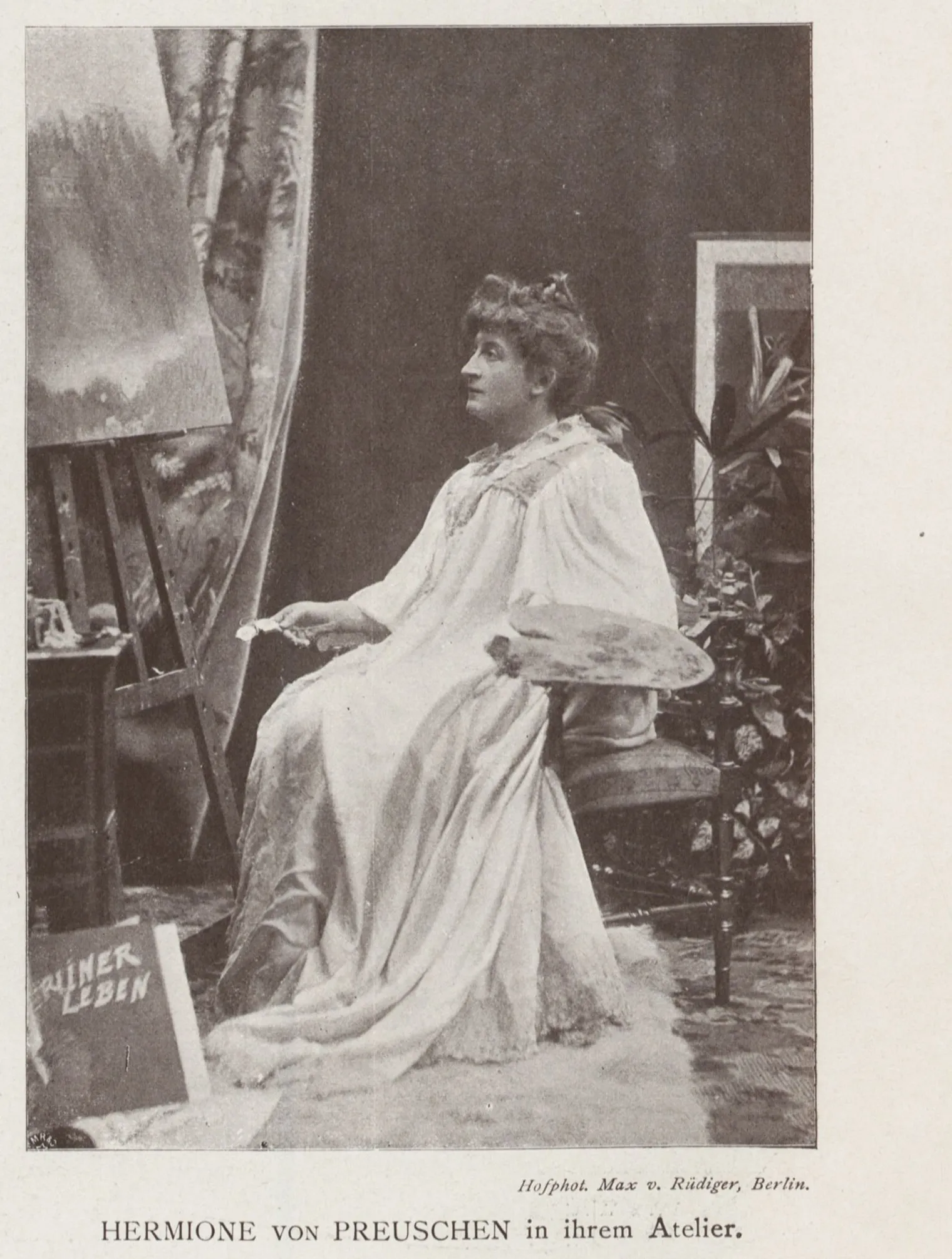 The painter seated at an easel in a loose pale gown, brush extended towards the canvas, an oval palette resting beside her, a large framed picture and palms behind, a magazine propped against the easel.
