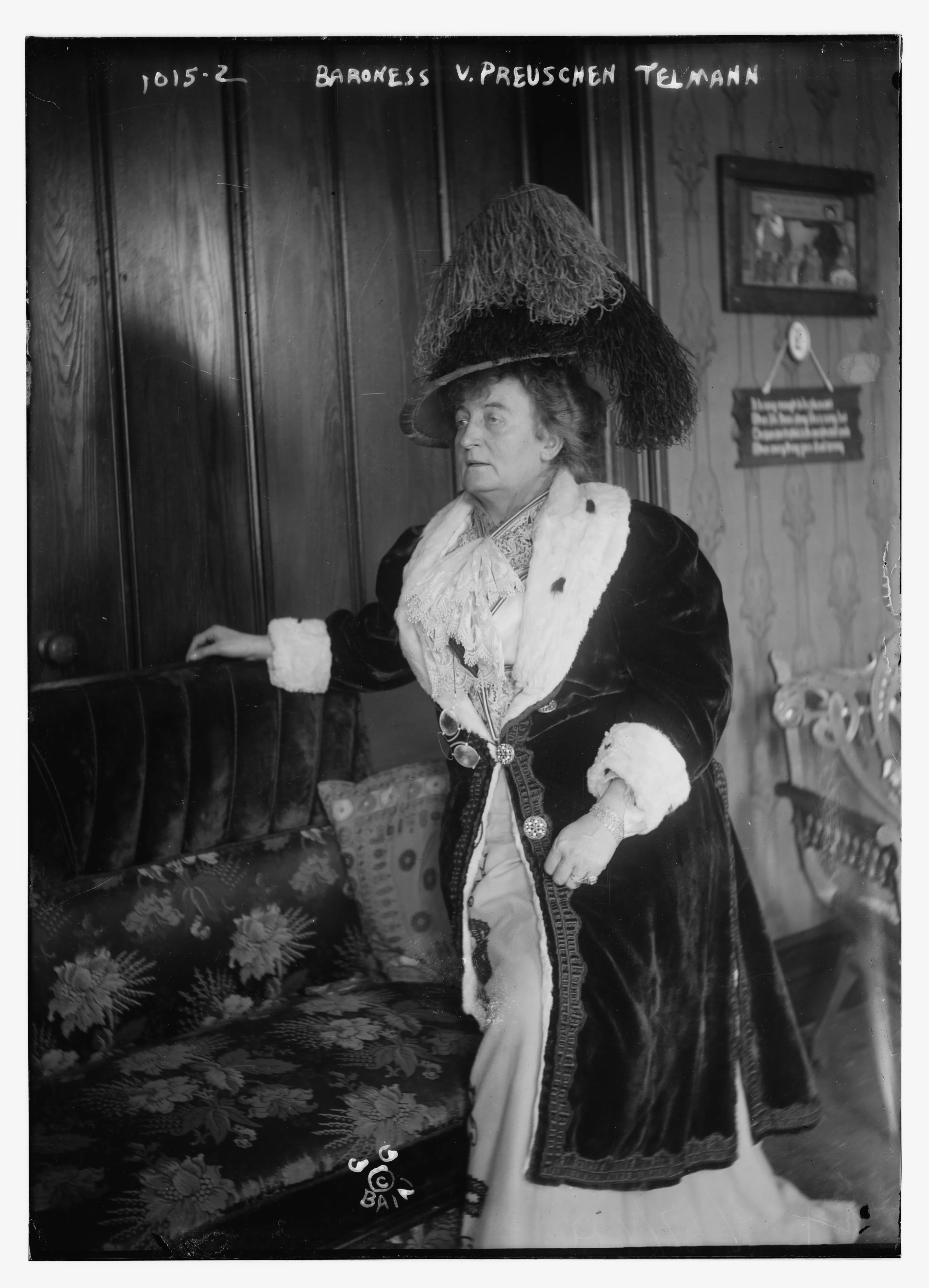 A woman in an enormous plumed hat and a fur-trimmed velvet coat stands with one hand resting on the back of a sofa, in a panelled interior hung with pictures.