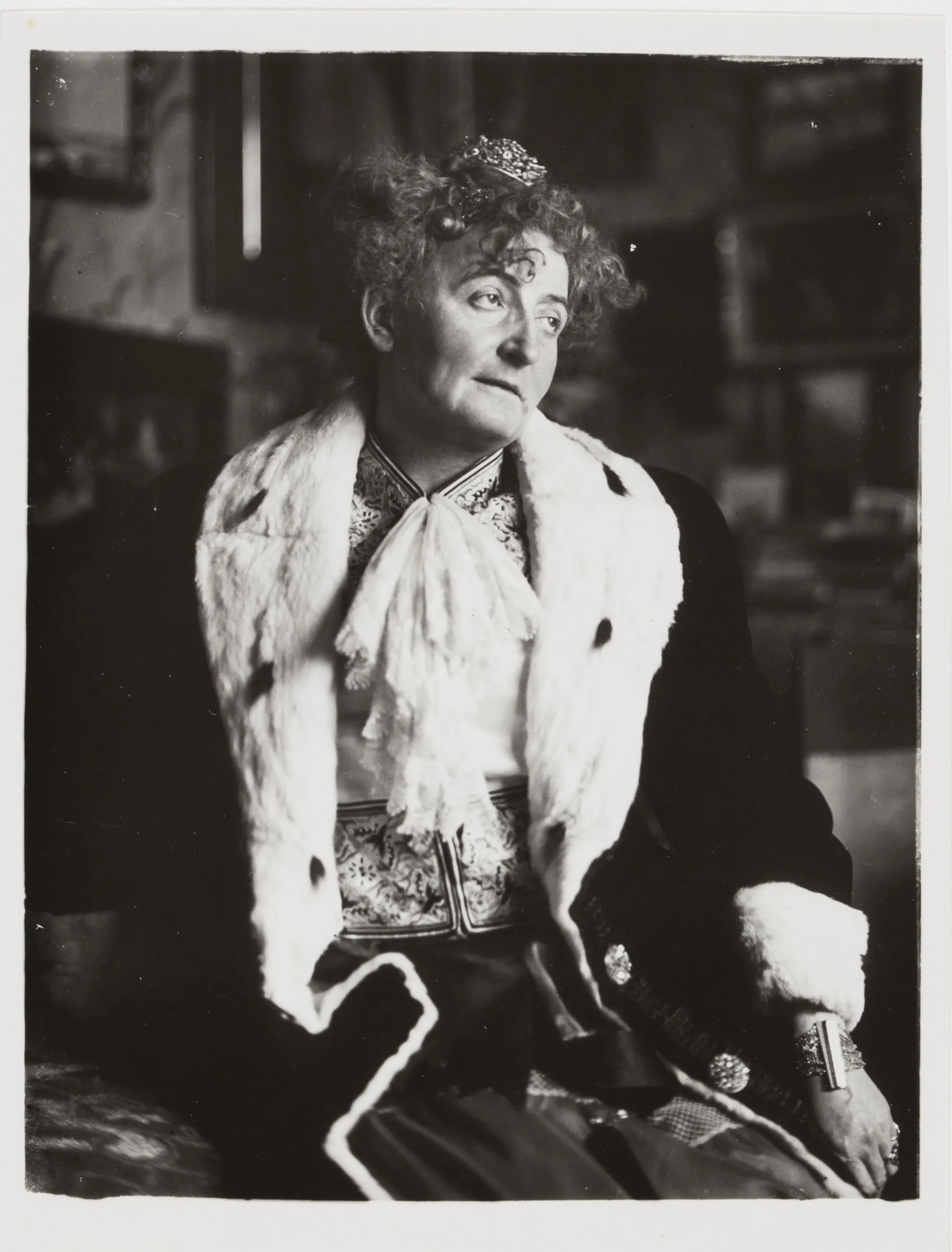 An older woman in a fur-trimmed coat over an embroidered jacket, an ornament in her hair, seated before a wall of framed pictures and looking away from the camera.