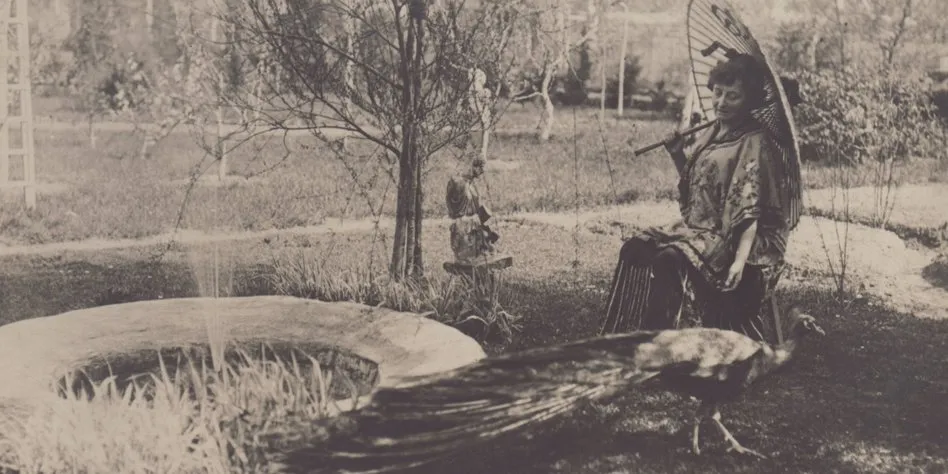 A woman in a loose patterned robe sits in a young garden under a painted paper parasol; a peacock walks across the grass in front of her, beside a low stone basin.